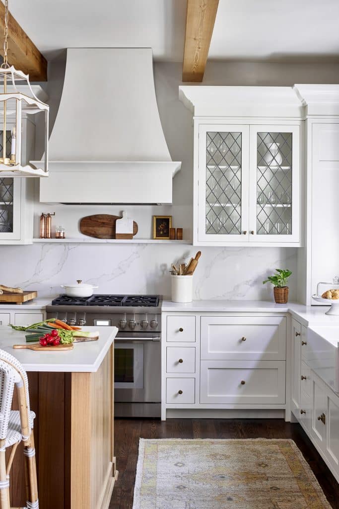 Transitional kitchen with white cabinetry, leaded glass cabinet doors, natural wood ceiling beams, and white range hood.