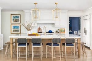 Transitional coastal style kitchen with large natural stained kitchen island, white granite counter, large brass pendant lights and natural stained floor.