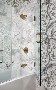 Marble tile shower with glass door, brass hardware, and gray and white floral wallpaper.