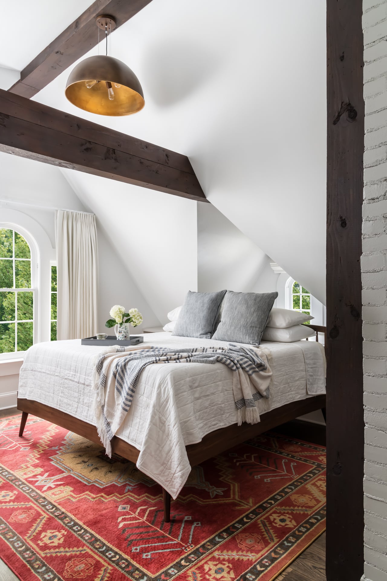 Transitional attic bedroom with natural wood exposed beams, vaulted ceiling, and brass dome pendant light.