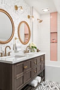 Children's bathroom with natural stained double sink vanity, round brass mirrors, simple cat patterned wallpaper, pink and white shower tile, and geometric floor tile.